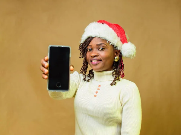 An African lady pointing a smart phone to the camera and also having a Christmas cap on her head