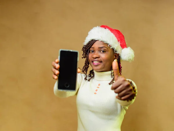An African lady pointing a smart phone to the camera, doing thumbs up gesture and also having a Christmas cap on her head