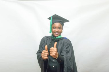 Stock photo of an African female graduand or graduating students in her black graduation outfit and joyfully doing thumbs up gesture