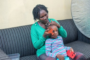 Afrikalı kadın anne, küçük kızıyla birlikte oturuyor, taraklı saçlarını siyah bir kanepede, akıllı bir telefonla kızın ellerinde yapıyor.