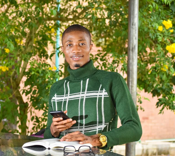 A light skinned african guy holding a smart phone and wearing a turtle-neck polo sits with a book in front of him during leisure time