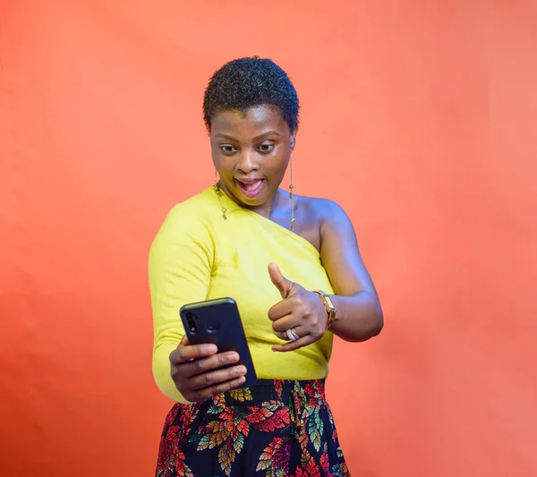 A female African Nigerian looking into a smart phone in her hands, doing thumbs up gesture and wearing a yellow top with a low cut hairstyle on her head