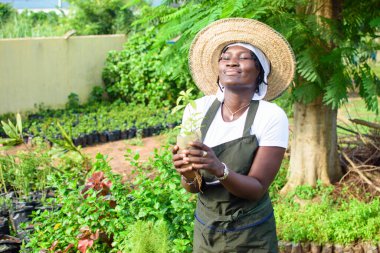 Happy African female gardener, florist or horticulturist wearing an apron and a hat, holding a bag of plant as she works in a green and colorful flowers garden