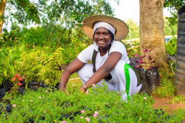 African female gardener, florist or horticulturist wearing an apron and a hat, working while squatting in a green and colorful flowers and plants garden clipart