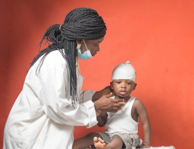  Burun maskesi ve steteskobu olan Afrikalı hemşire, doktor ya da tıp uzmanı. Kafasına sarılmış yara bandı olan küçük bir çocuk üzerinde sağlık kontrolü ve tedavi yapıyor. 