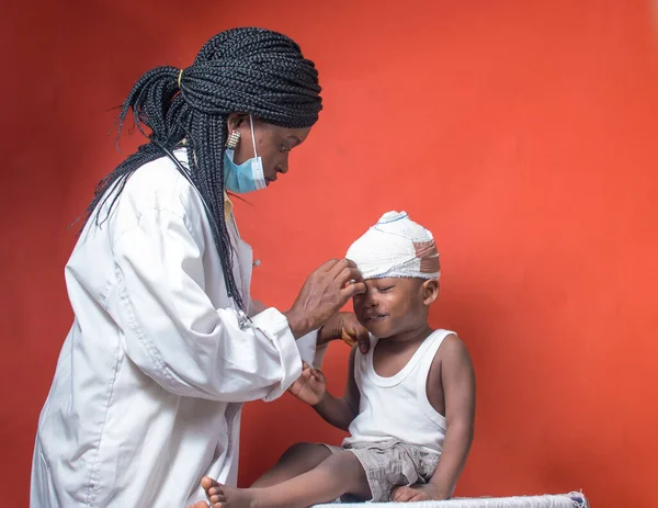  Burun maskesi ve steteskobu olan Afrikalı hemşire, doktor ya da tıp uzmanı. Kafasına sarılmış yara bandı olan küçük bir çocuk üzerinde sağlık kontrolü ve tedavi yapıyor. 