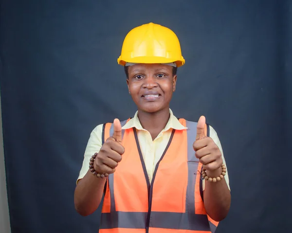 African Nigerian female construction or civil engineer, architect or builder with yellow safety helmet and orange reflective jacket, doing thumbs up gestures