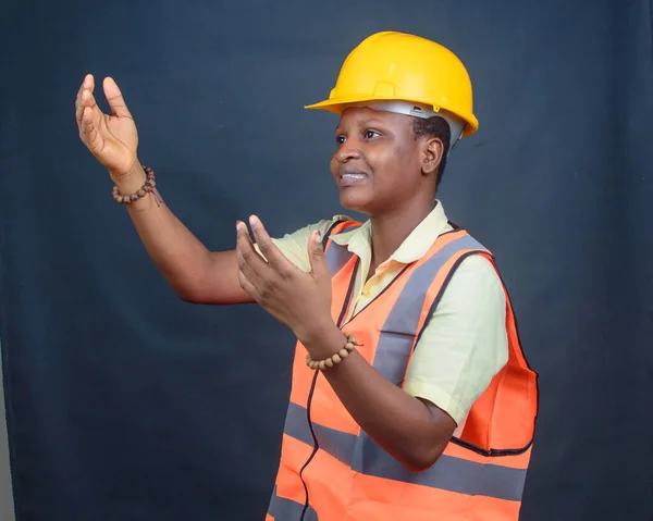 African Nigerian female construction or civil engineer, architect or builder with yellow safety helmet and orange reflective jacket, beckoning or signaling to someone