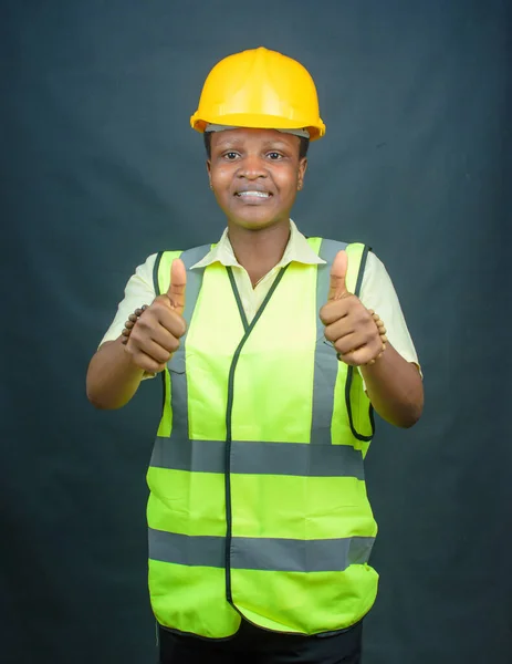 African Nigerian female construction or civil engineer, architect or builder with yellow safety helmet and green reflective jacket, doing thumbs up gestures