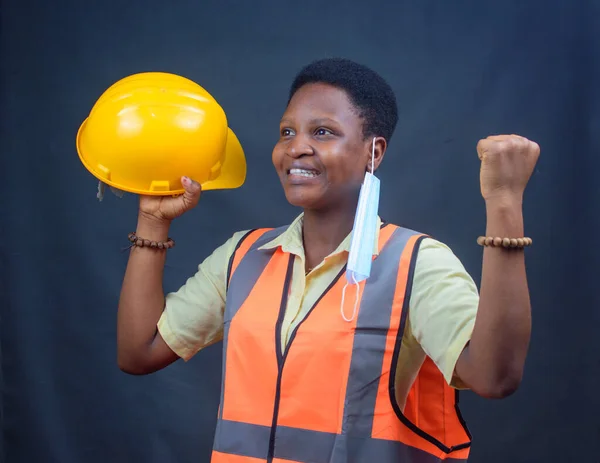 Excited African Nigerian female construction or civil engineer, architect or builder holding a yellow safety helmet and wearing a orange reflective jacket