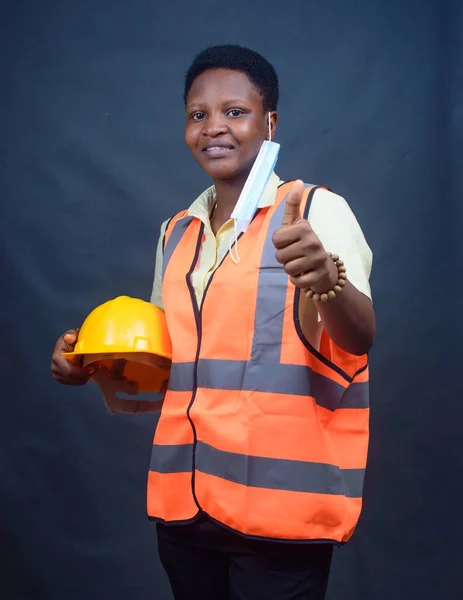African Nigerian female construction or civil engineer, architect or builder with yellow safety helmet and orange reflective jacket, doing thumbs up gestures