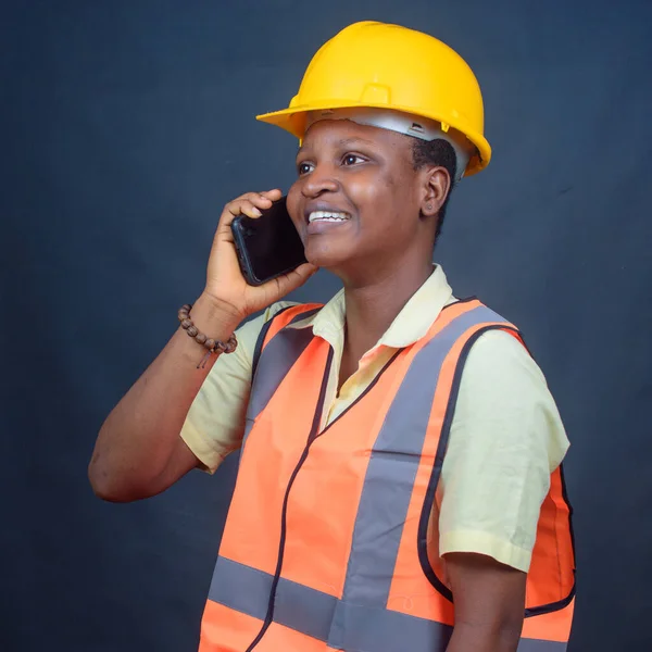 An african Nigerian female construction or civil engineer, architect or builder with orange safety helmet, making calls with a smart phone