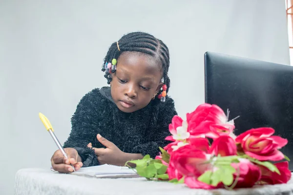 Little cute african girl child with an afro hair style studying as she writes in a book with a flowers and a black laptop in her front