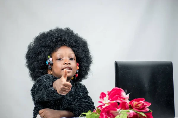 Smiling little African girl child with an afro hair style giving thumbs up gesture with a laptop and flowers in front of her 