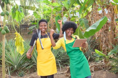 Two excited African female farmers or business women with colorful apron, holding farming tool and a tablet on a farmland