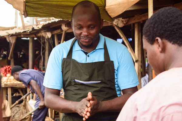 stock photo of male African grocery seller or business man with apron, standing at his stall in a market, ready to sell to customers