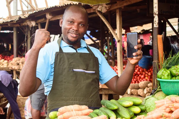 stock photo of a happy male African grocery seller with apron and smart phone, ready to sell to customers
