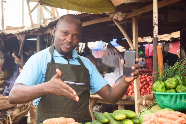 stock photo of a happy male African grocery seller with apron and smart phone, ready to sell to customers