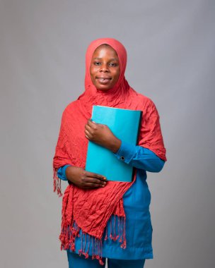 An African Muslim lady, female student or businesswoman with head scarf called hijab, holding a book to her chest while standing