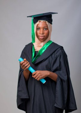 African Nigerian Muslim lady, student or graduand, wearing an hijab head scarf and graduation outfit, with a wrapped book or certificate in her hands