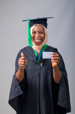 African Nigerian Muslim lady, student or graduand, wearing an hijab head scarf and graduation gown, holding a small white card and doing thumbs up gestures with her second hand
