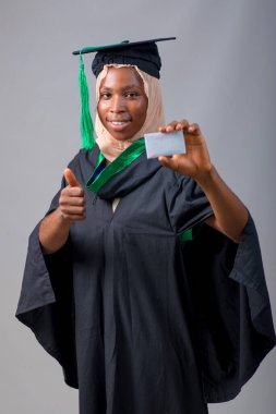 African Nigerian Muslim lady, student or graduand, wearing an hijab head scarf and graduation gown, holding a small white card and doing thumbs up gestures with her second hand