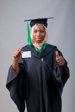 African Nigerian Muslim lady, student or graduand, wearing an hijab head scarf and graduation gown, holding a small white card and doing thumbs up gestures with her second hand