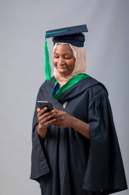 African Nigerian Muslim lady, student or graduand, wearing an hijab head scarf and graduation gown and also smile as she look at a smart phone in her hand