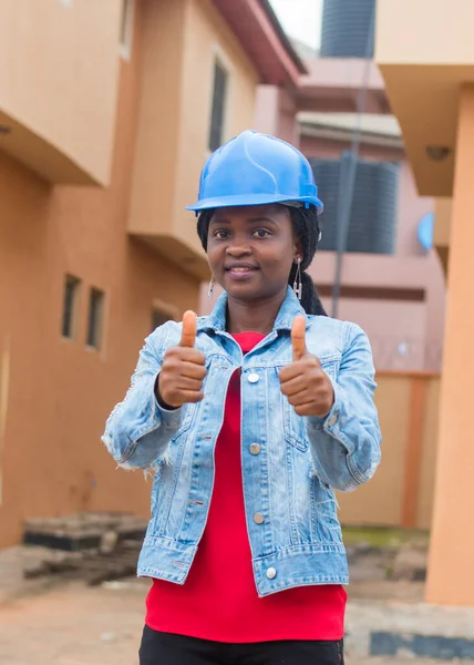 African lady or woman from Nigeria with blue safety helmet, doing thumbs up gestures as she represents industrial professionals like construction specialist, builders, architect and engineer