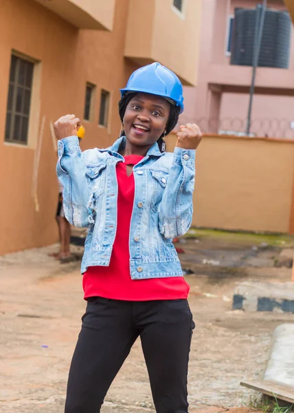 Happy African lady or woman from Nigeria with blue safety helmet, dancing and celebrating as she represents industrial professionals like construction specialist, builders, architect and engineer