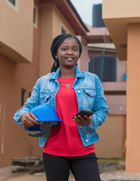 Portrait of a cute African lady or woman from Nigeria, holding a phone and blue safety helmet as she represent industrial professionals like construction specialist, builders, architect and engineer