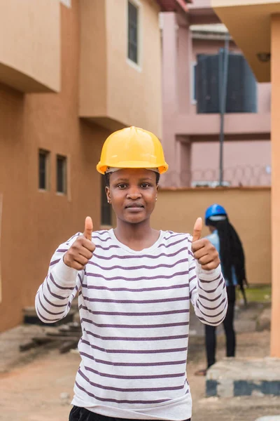 An African lady or woman from Nigeria with yellow safety helmet, doing thumbs up gesture as she represents industrial professionals like construction specialist, builders, architect and engineer