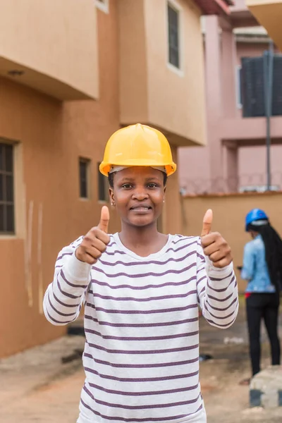 An African lady or woman from Nigeria with yellow safety helmet, doing thumbs up gesture as she represents industrial professionals like construction specialist, builders, architect and engineer