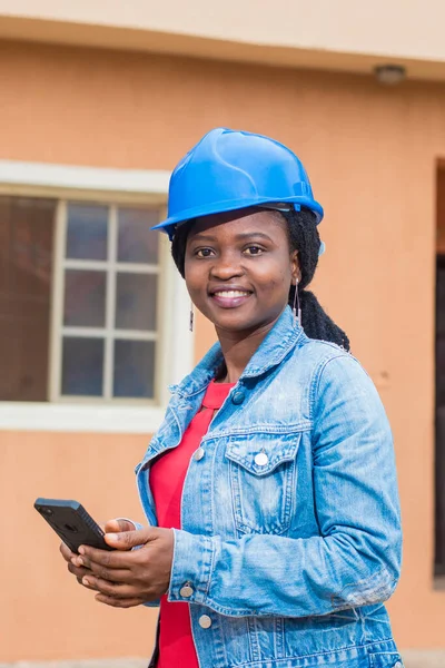 African lady or woman from Nigeria with blue safety helmet, holding a phone as she represents industrial professionals like construction specialist, builders, architect and engineer