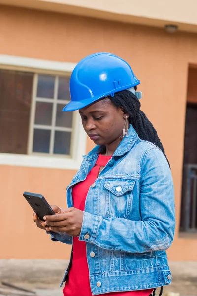African lady or woman from Nigeria with blue safety helmet, holding and using a phone as she represents industrial professionals like construction specialist, builders, architect and engineer