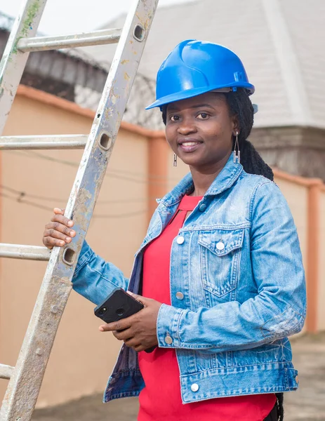 African lady or woman from Nigeria with blue safety helmet, holding a ladder and a phone as she represents industrial professionals like construction specialist, builders, architect and engineer