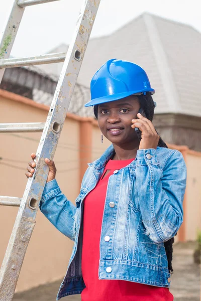 African lady or woman from Nigeria with blue safety helmet, making call while holding a ladder as she represents industrial professionals like construction specialist, builders, architect and engineer