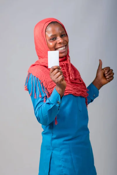 An African Muslim lady with head scarf, cover or hijab, holding a small white card and doing thumbs up gesture while standing