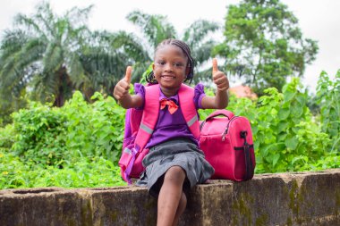 An African girl child in school uniform and with a backpack, doing thumbs up gestures and sitting outside on a low fence