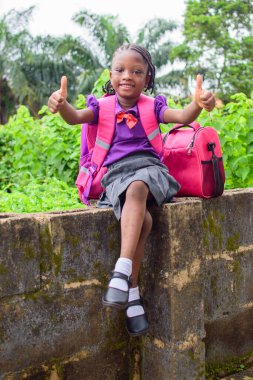 An African girl child in school uniform and with a backpack, doing thumbs up gestures and sitting outside on a low fence