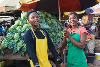 Two happy African business women or female traders wearing colorful aprons and standing together in front of their vegetable stall in a market place