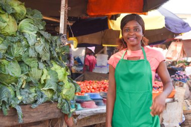 Happy African business woman or female trader wearing a green apron and standing at her stall in a market