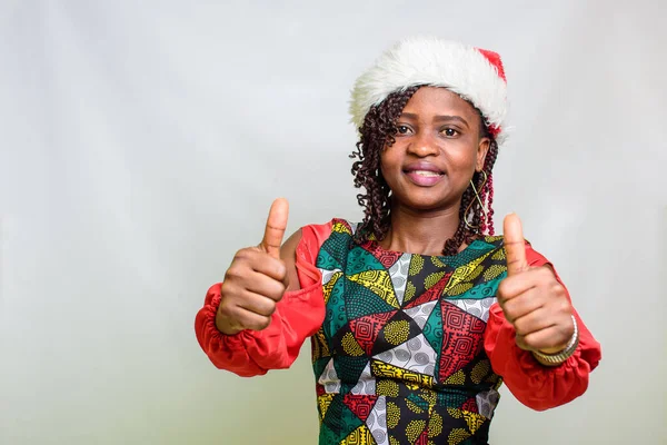 An african lady from Nigeria doing thumbs up gestures with both hands while having a christmas cap on her head