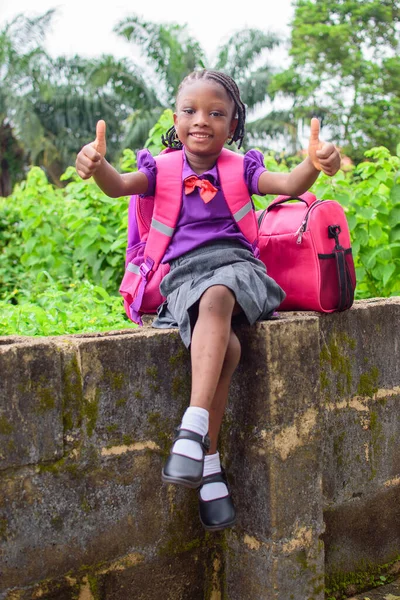 An African girl child in school uniform and with a backpack, doing thumbs up gestures and sitting outside on a low fence