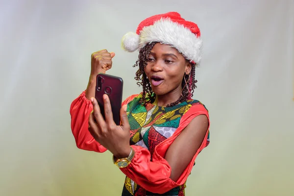 A happy african lady from Nigeria looking into the smart phone in her hands and also wearing a christmas cap