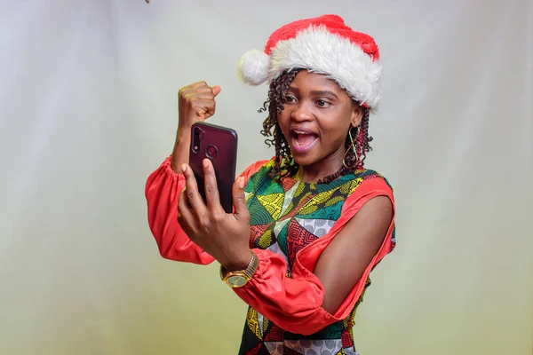A happy african lady from Nigeria looking into the smart phone in her hands and also wearing a christmas cap