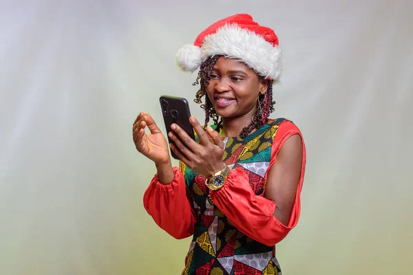 An african lady from Nigeria looking into the smart phone in her hands and also wearing a christmas cap