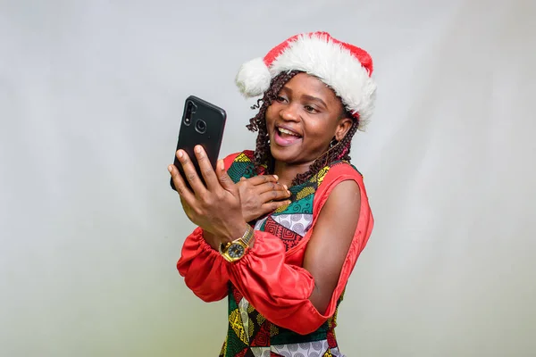 An african lady from Nigeria looking into the smart phone in her hands and also wearing a christmas cap