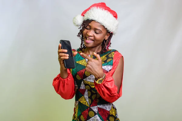 An african lady from Nigeria pointing to the smart phone in her hands and also wearing a christmas cap