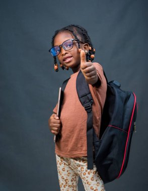 A female African girl child or kid student from Nigeria happily doing thumbs up to the camera while carrying an educational smart tablet in her hands and a school back on her back